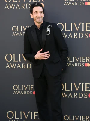 Adrien Brody smiling on the Olivier Awards red carpet, dressed in a black Dior tuxedo with peak lapels and a black wing-tip collar shirt, accessorized with black leather derbies.