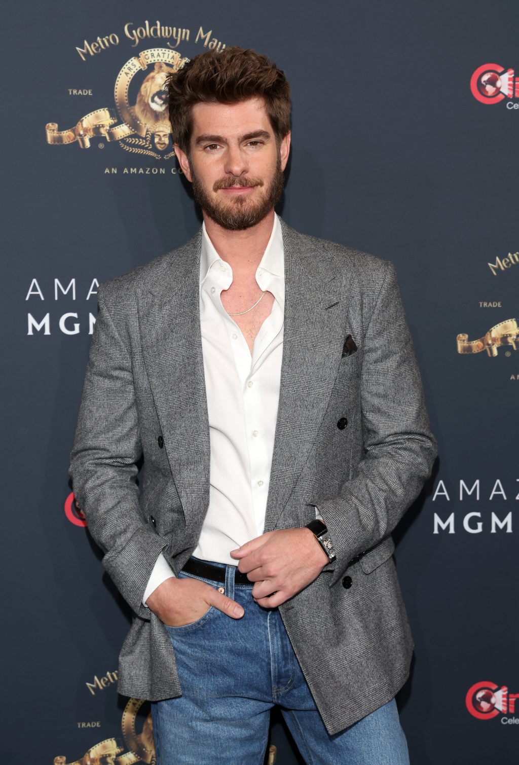 Andrew Garfield poses solo on the red carpet in a grey Dunhill blazer, white shirt, and blue jeans at the CinemaCon 2025 event.