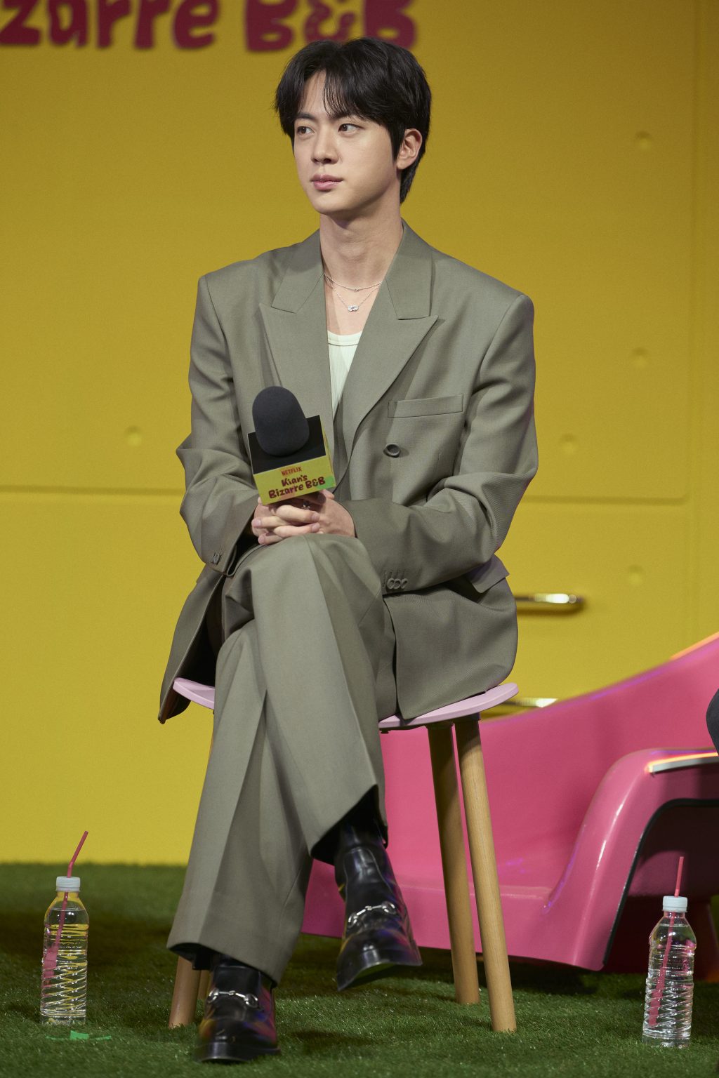 Jin sits on a chair at a press conference, wearing a sage green Gucci suit with a mint inner top and black leather boots.