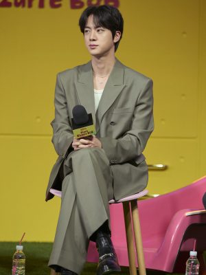 Jin sits on a chair at a press conference, wearing a sage green Gucci suit with a mint inner top and black leather boots.