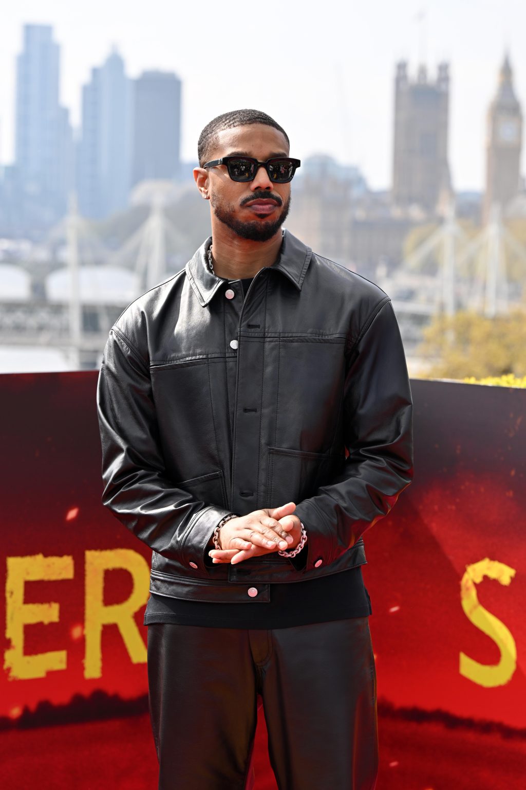 Michael B. Jordan wearing a black leather jacket and pants by GapStudio, standing in front of a red and yellow backdrop with the London skyline behind him