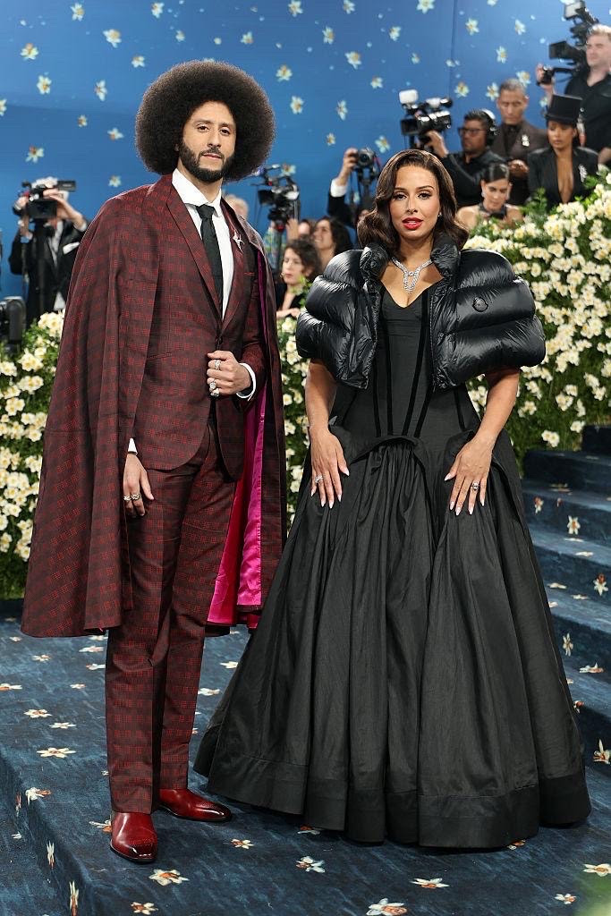 Colin Kaepernick wearing a red silk jacquard suit with a matching overcoat at the 2025 Met Gala, standing beside his partner on the floral carpeted stairs.