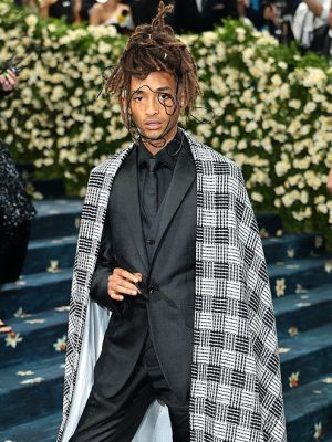 Jaden Smith wearing a custom black suit with wide-leg trousers and a houndstooth cape at the 2025 Met Gala, standing on the steps lined with floral carpet.