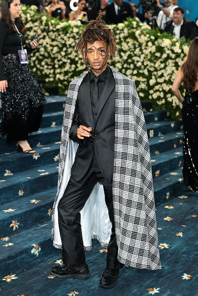 Jaden Smith wearing a custom black suit with wide-leg trousers and a houndstooth cape at the 2025 Met Gala, standing on the steps lined with floral carpet.