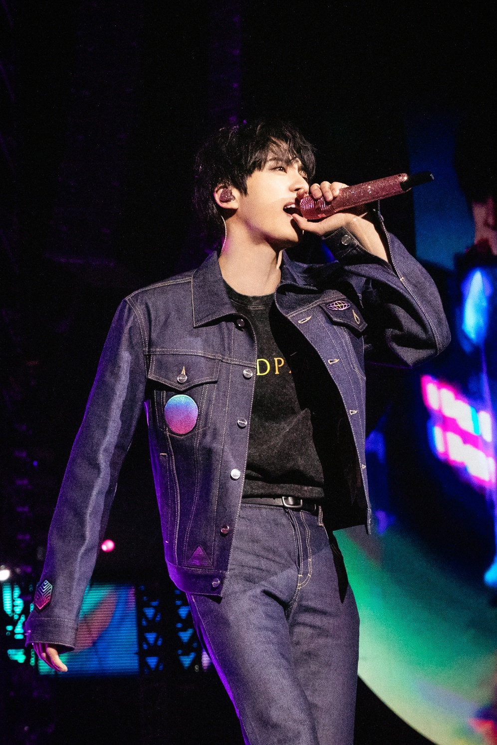 Jin of BTS performing in a dark denim Gucci outfit, holding a pink microphone during Coldplay’s Music Of The Spheres concert.