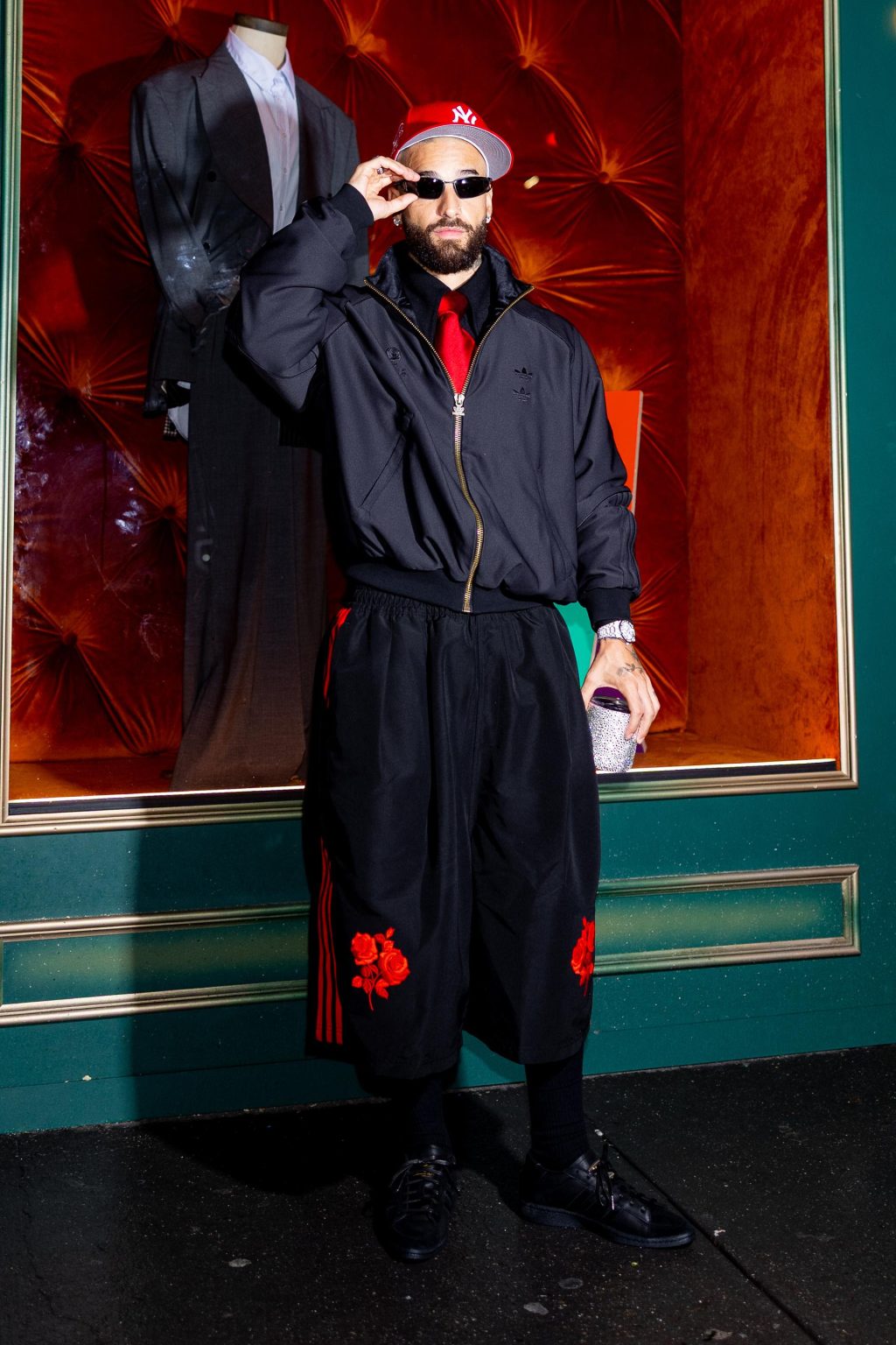 Maluma standing in front of a store window wearing adidas Originals x Willy Chavarria black jacket and long shorts with red accents, accessorized with a red Yankees cap and sunglasses.