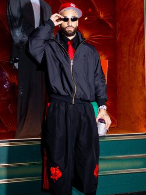 Maluma standing in front of a store window wearing adidas Originals x Willy Chavarria black jacket and long shorts with red accents, accessorized with a red Yankees cap and sunglasses.